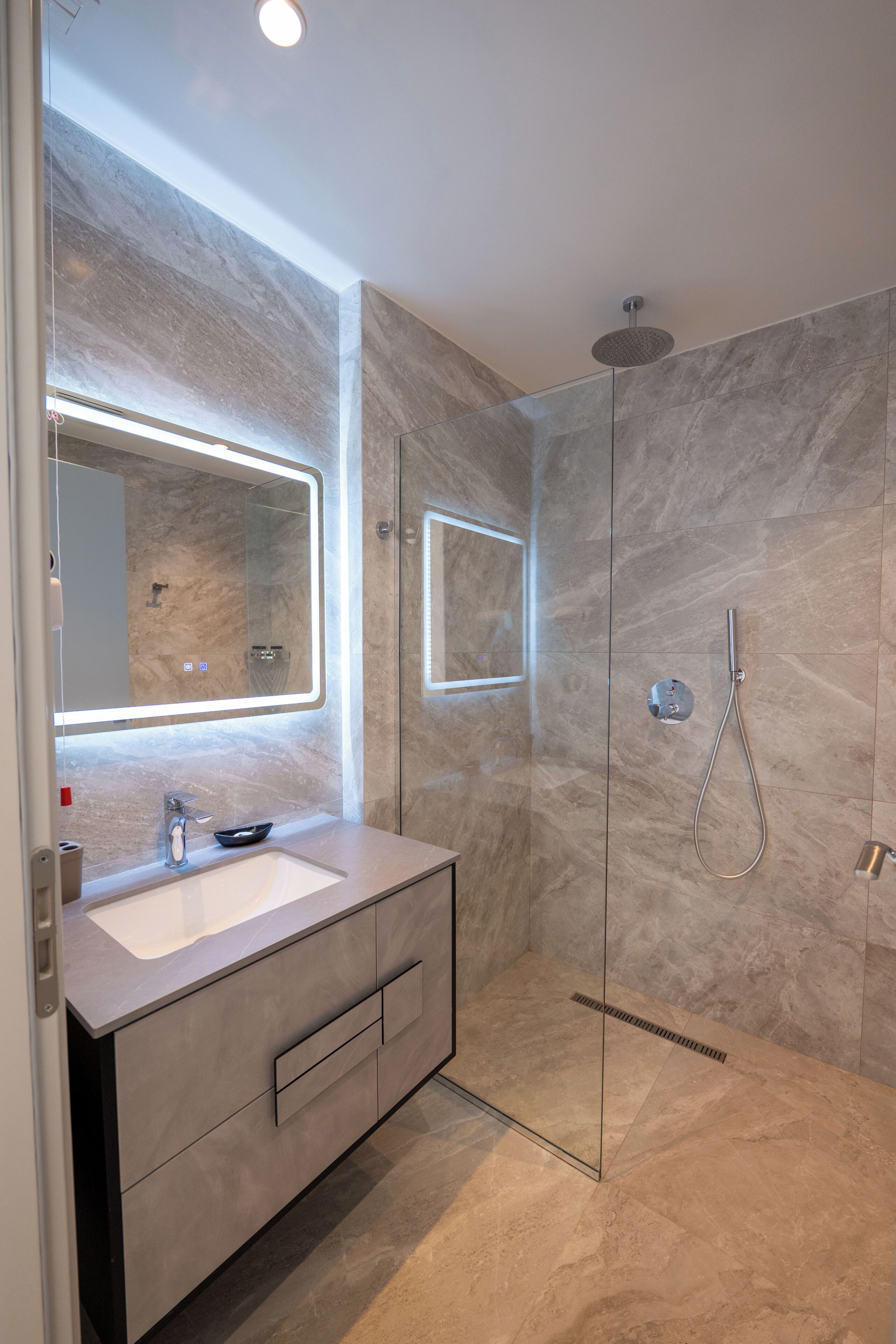 A modern bathroom with a glass-enclosed shower, a large mirror, and a marble-tiled wall.