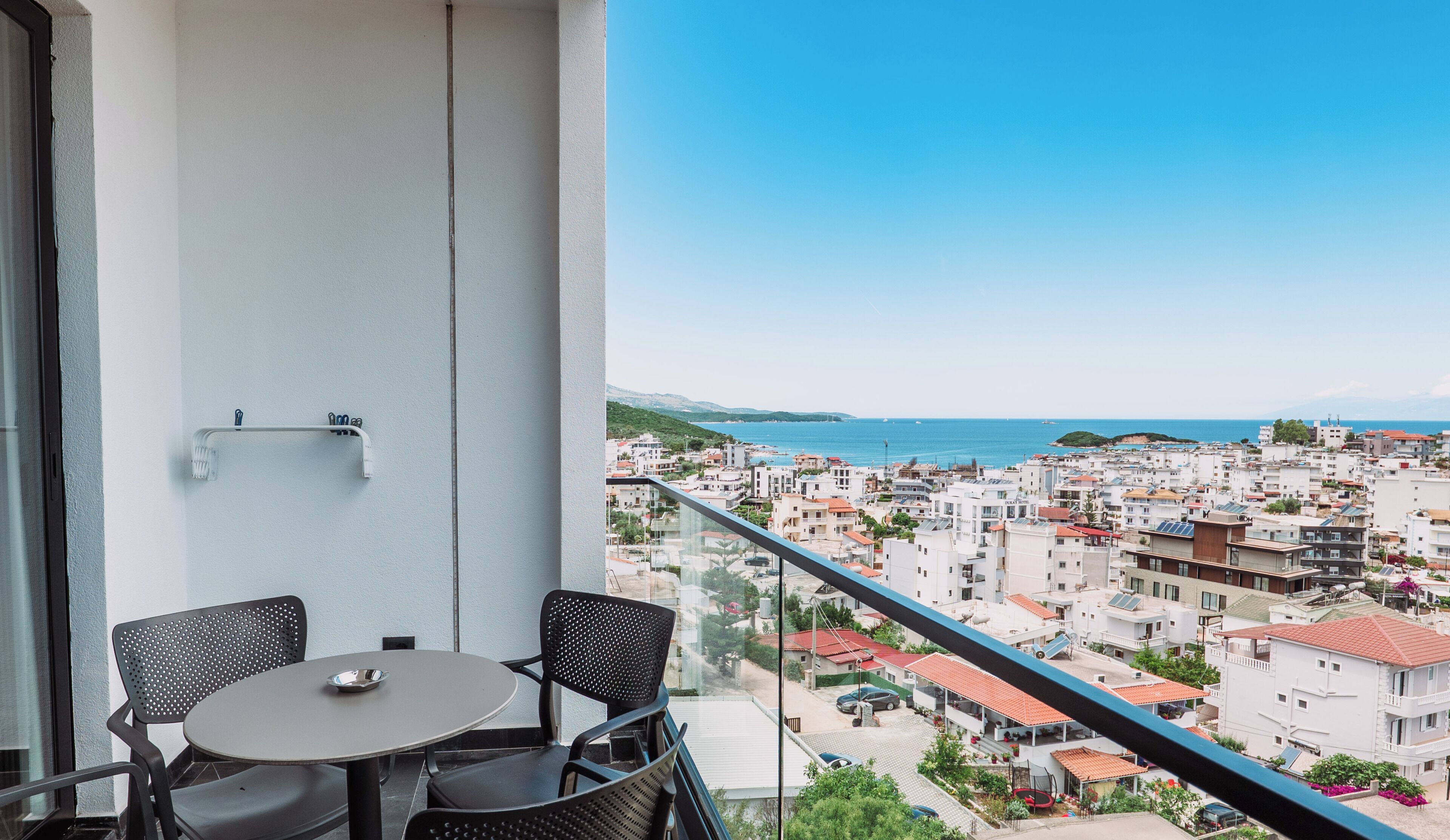 A balcony with a table and chairs, overlooking a coastal town with buildings and the sea.
