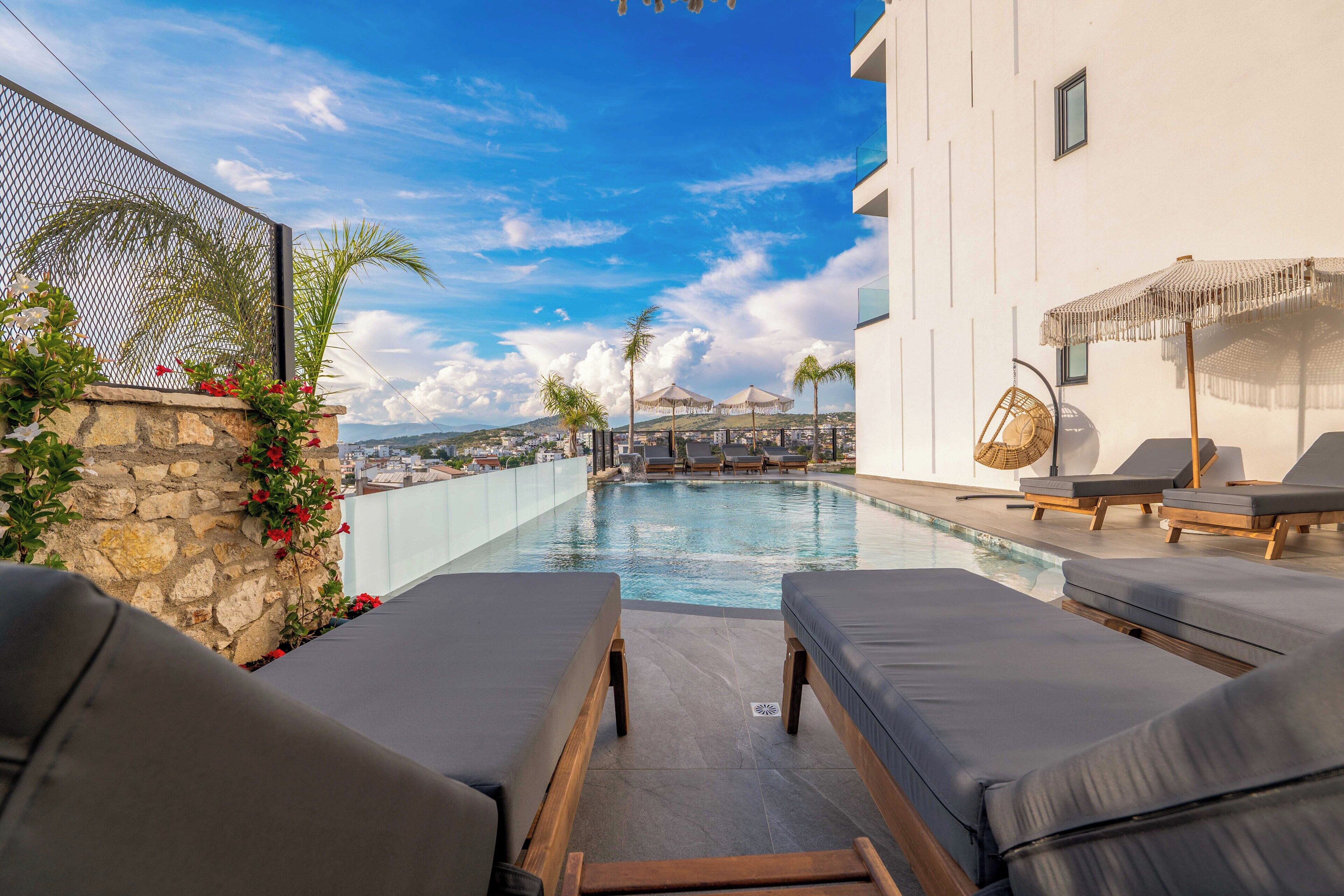 A rooftop pool area with lounge chairs, a stone wall with red flowers, and a view of the cityscape and mountains in the background.