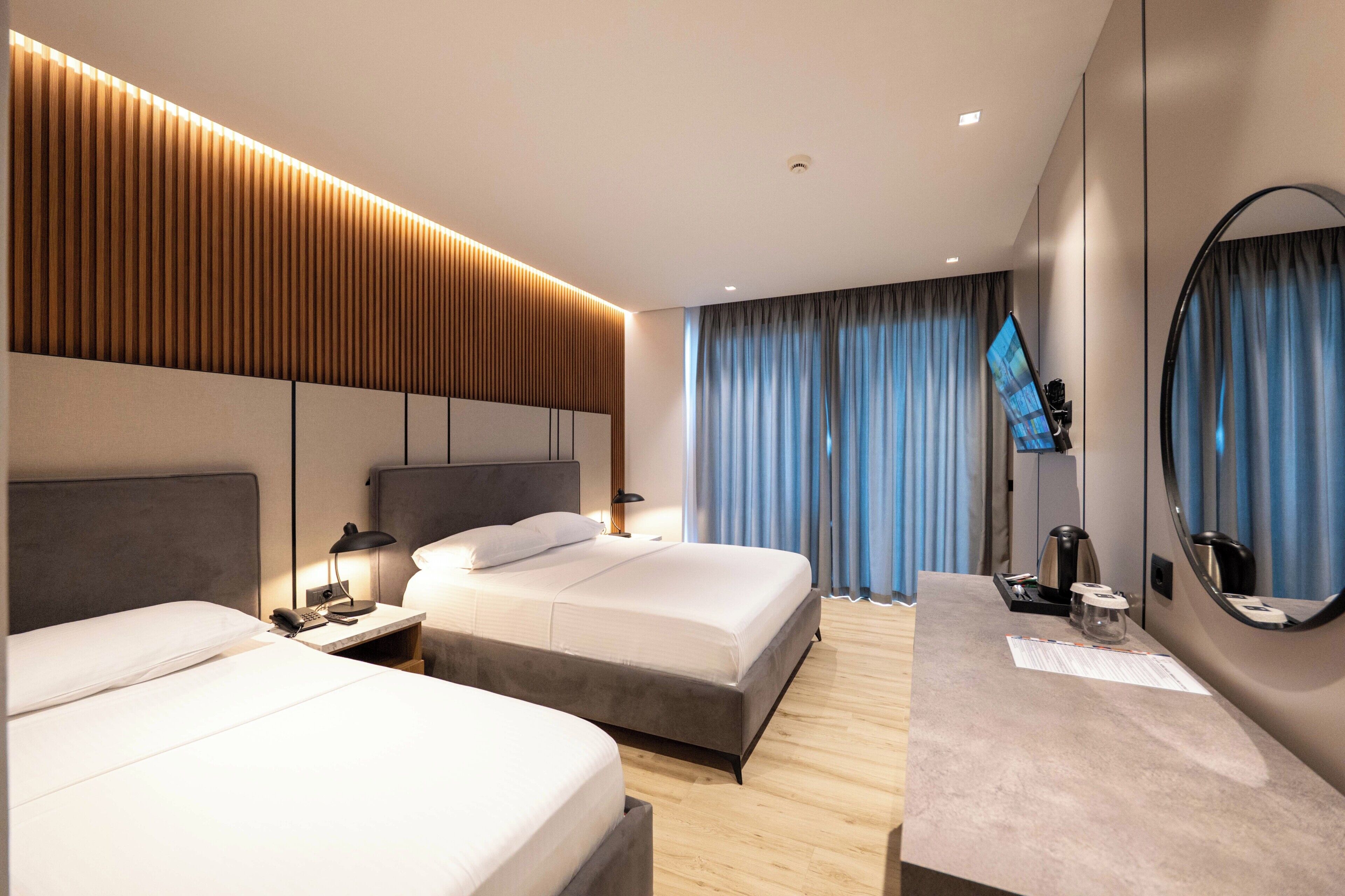 A modern hotel room with two beds, a desk, a mirror, and a television.