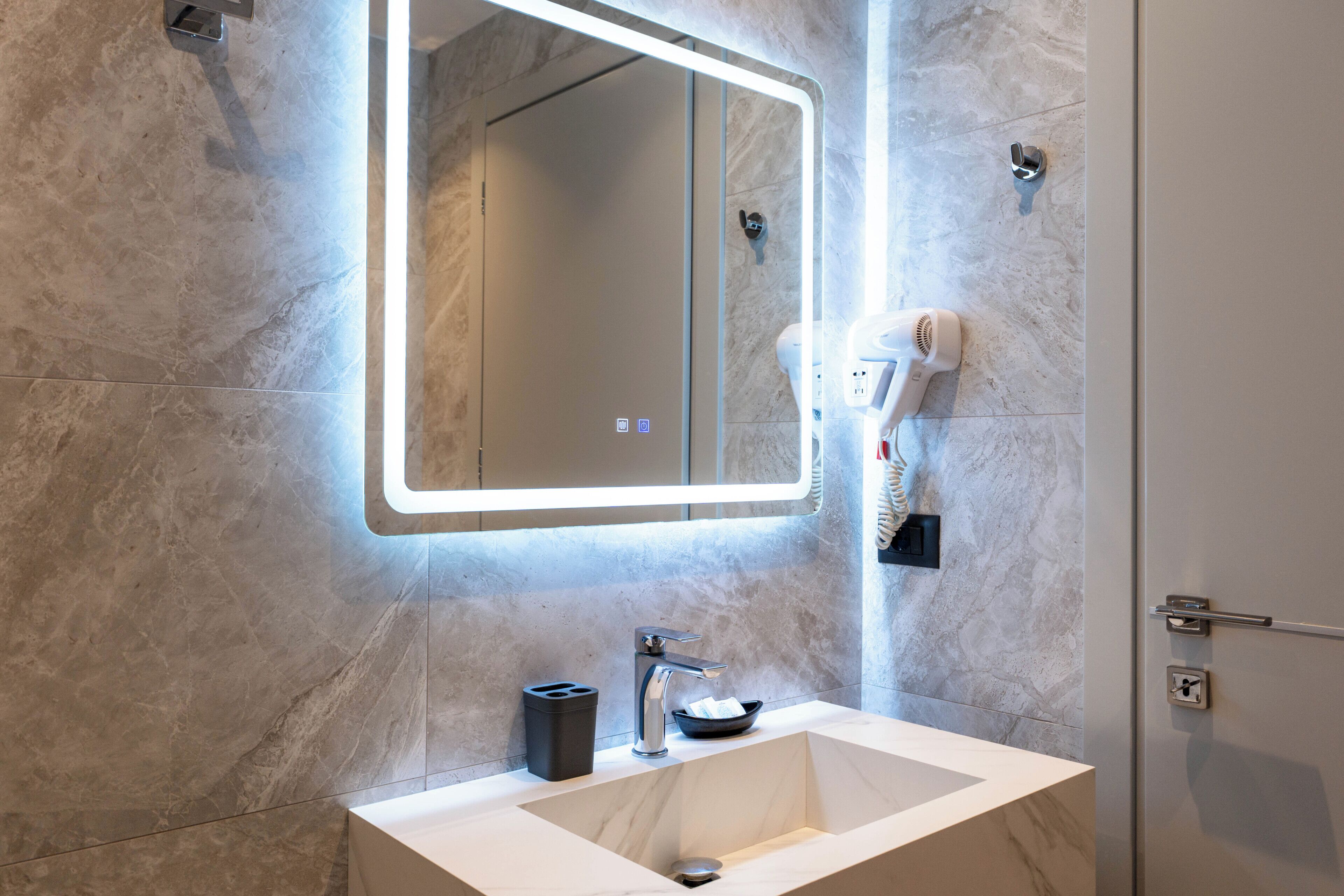 A bathroom with a marble sink, a mirror with integrated lighting, a hairdryer, and a towel rack.