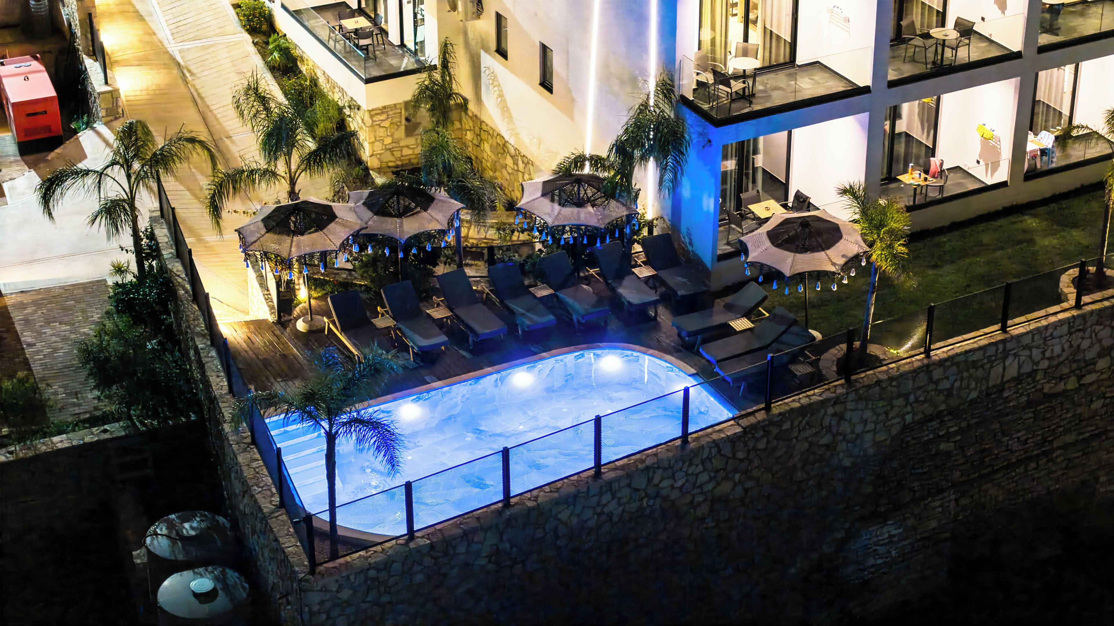 An outdoor swimming pool with lounge chairs and umbrellas, surrounded by stone walls and palm trees.