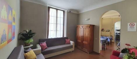 A living room with a sofa, a wooden wardrobe, a dining area, and a kitchen visible through an archway.