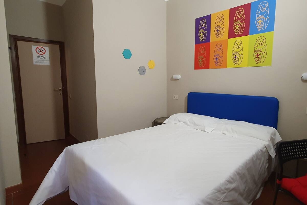 A bedroom with a bed, a blue headboard, a wall with a lion-themed artwork, a door with a no smoking sign, and a chair with a red cushion.