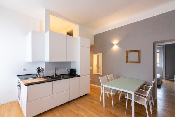 A compact kitchen with white cabinets, a black countertop, and a dining area with a green table and white chairs. - Sant'Ambrogio - Milan, central apartament for 5 (Milano)