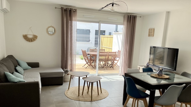 Ein modernes Wohnzimmer mit einer Couch, einem Esstisch und einem Fernseher. Eine Glastür verschiebt sich zu einem Außenbereich mit Tisch und Stühlen.