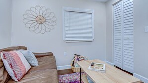 A room with a brown leather sofa, a wooden table with a magazine and a remote, a decorative wall piece, a window with blinds, and a white closet.
