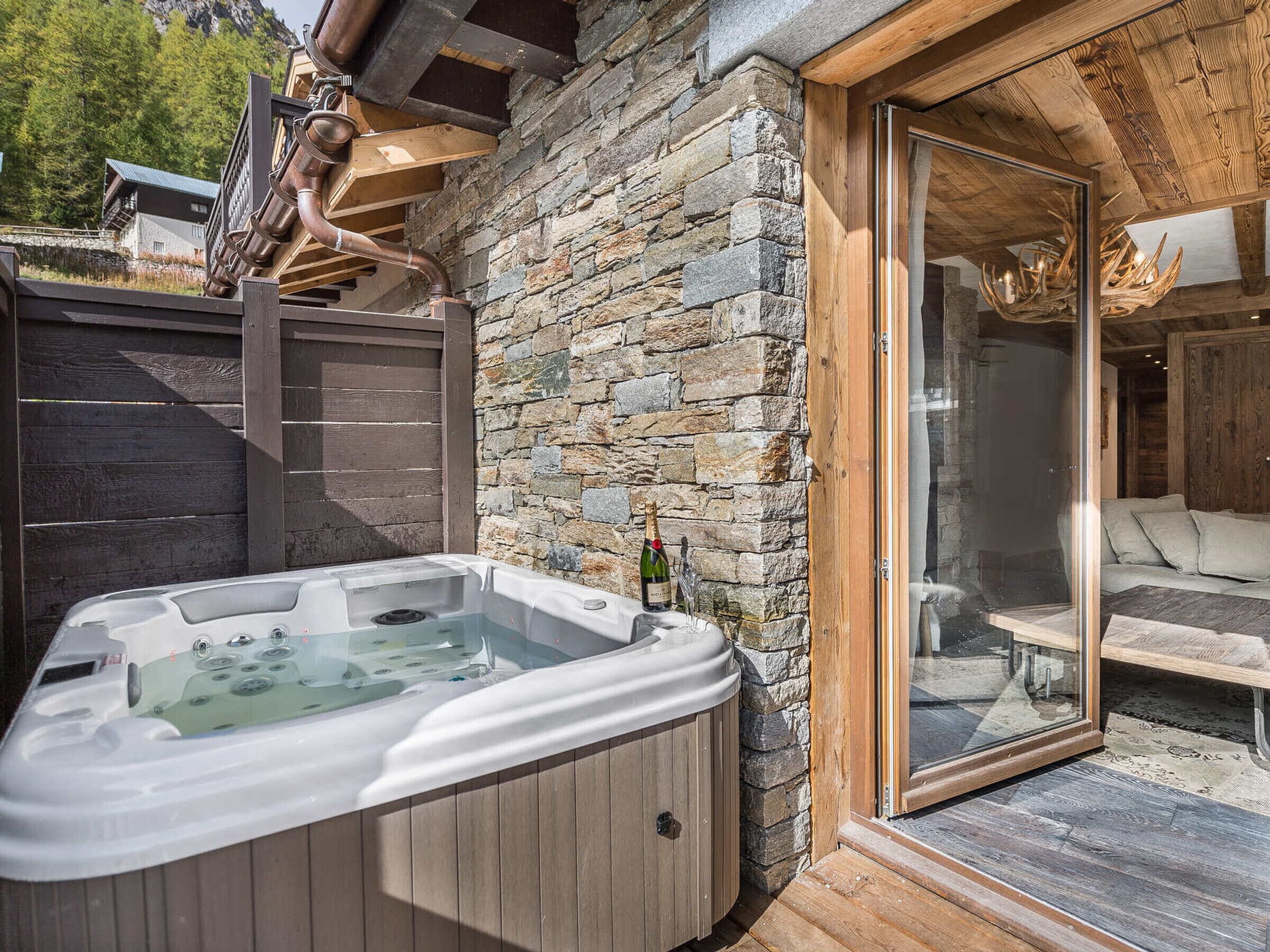 Een hottub buiten een stenen gebouw met een houten terras en een glazen deur die leidt naar een zithoek.