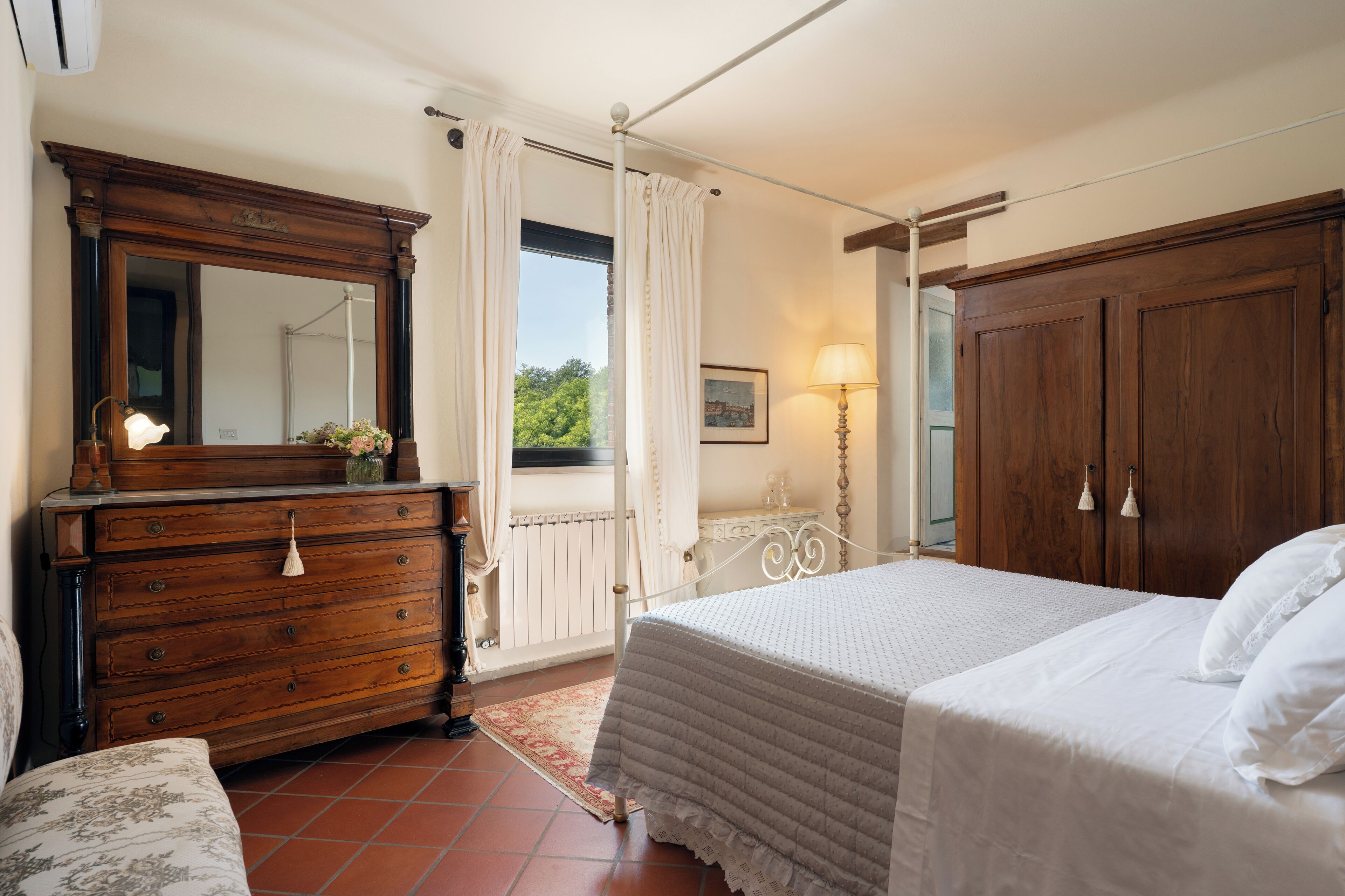 A bedroom with a wooden dresser, a large mirror, a bed with white bedding, a nightstand, and a window with curtains.