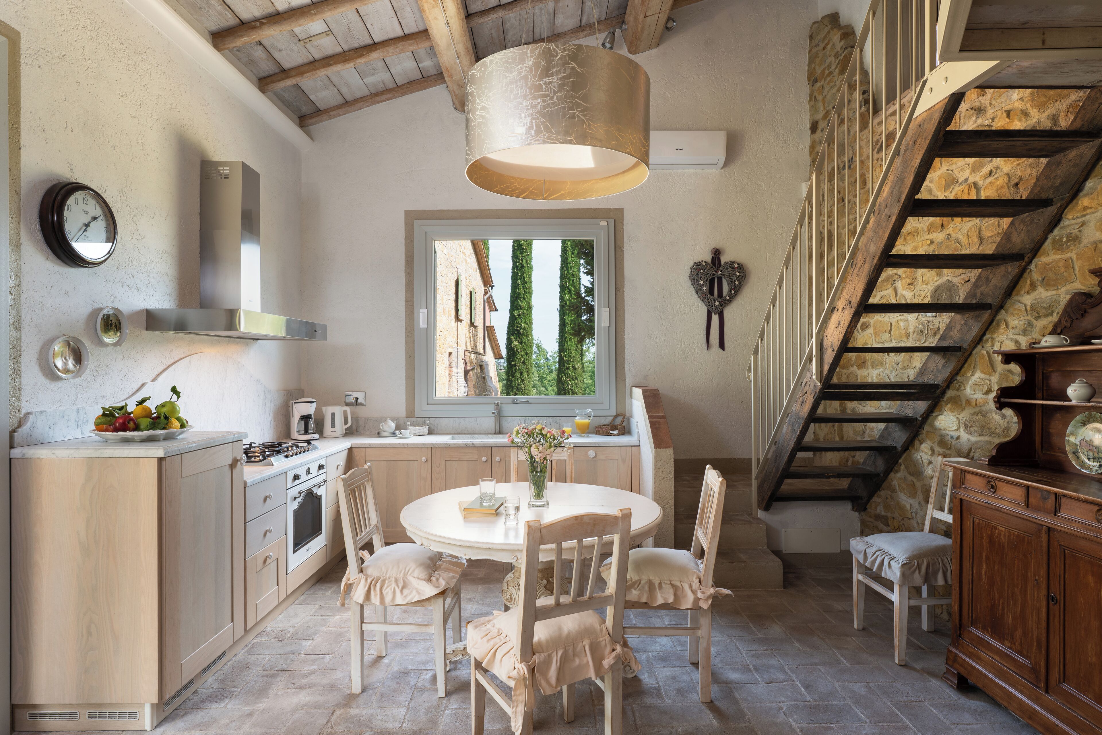 A kitchen with a round table, wooden chairs, a hanging light fixture, a stove, and a window with a view of trees.