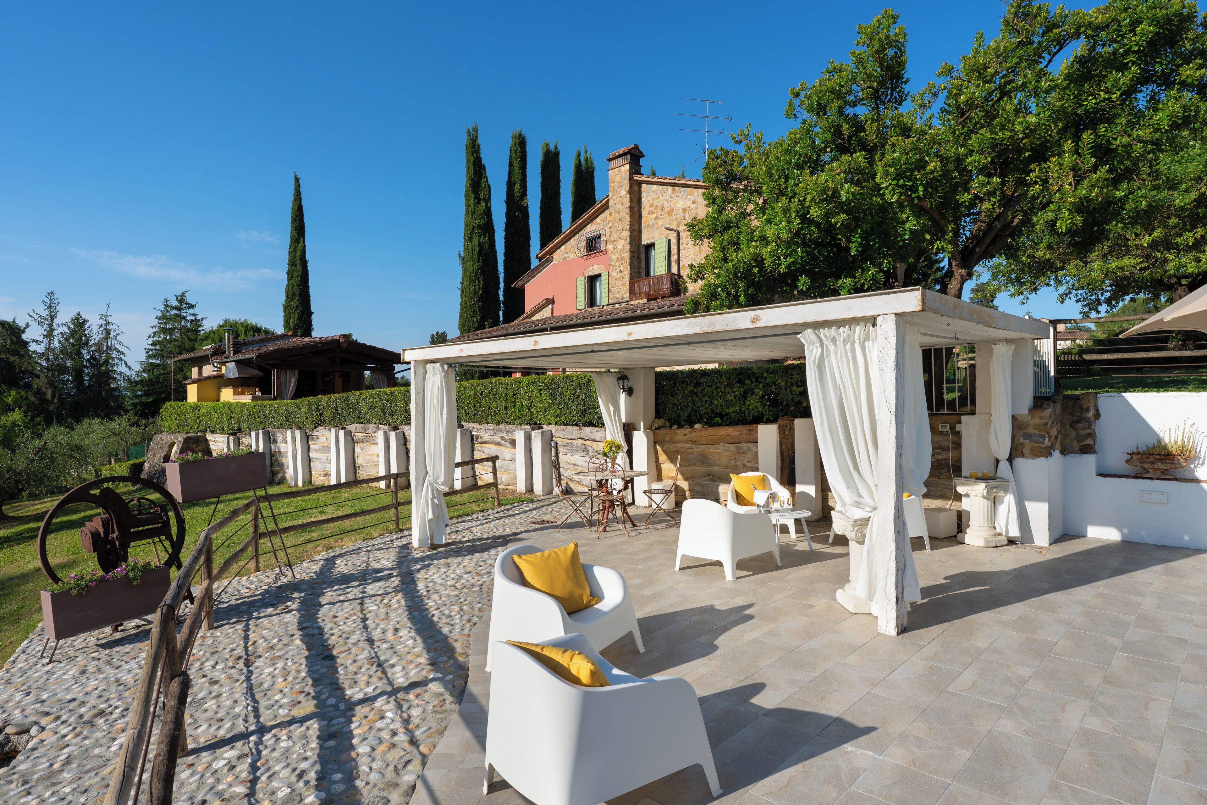 Une terrasse extérieure avec des meubles blancs, une pergola et un bâtiment en pierre en arrière-plan.