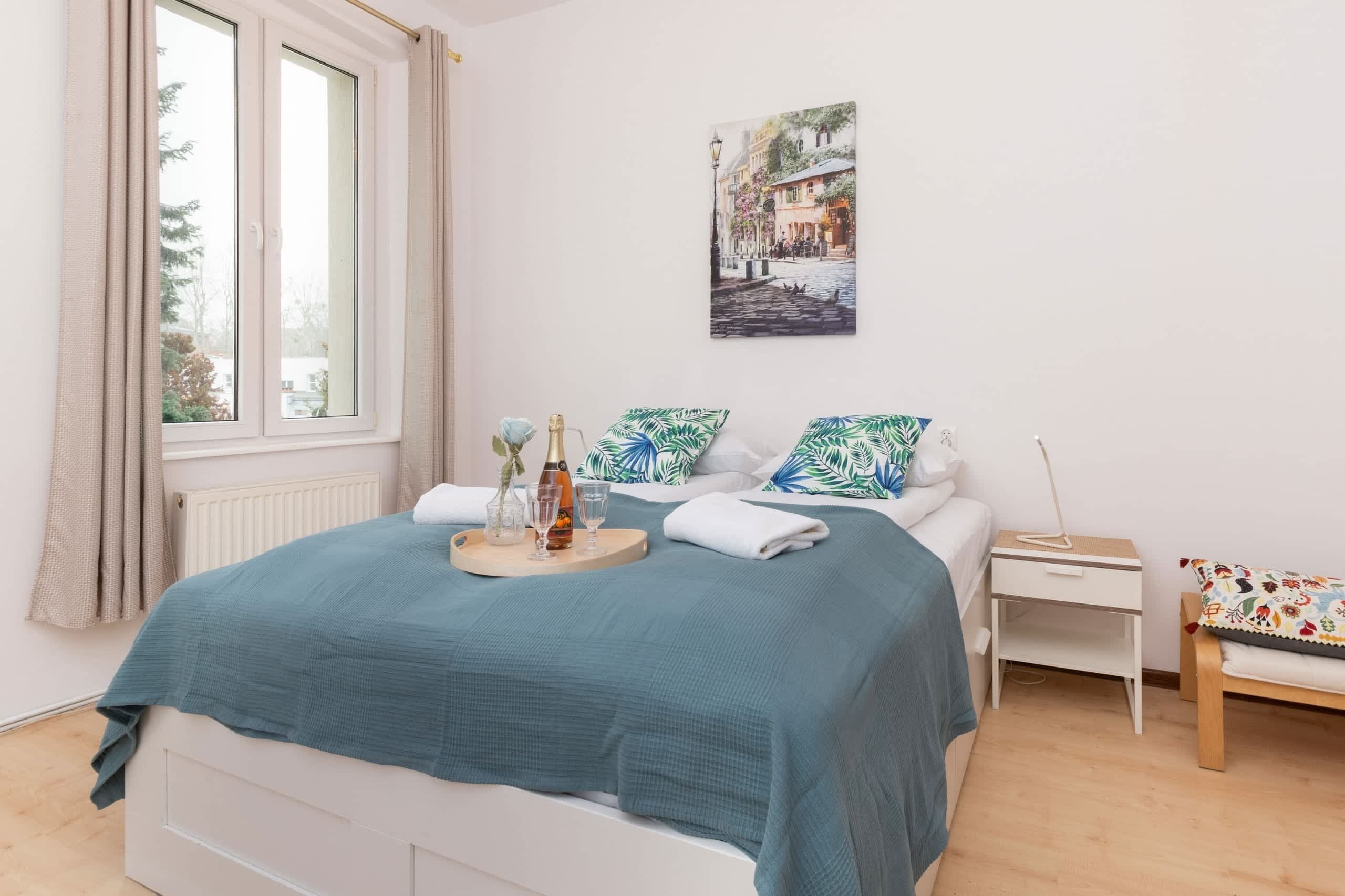 A neatly arranged bedroom with a bed, bedside tables, a nightstand, a window with curtains, and a wall-mounted picture.