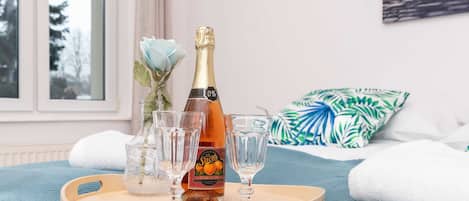 A bottle of champagne on a tray with glasses, a vase with a blue flower, and a tropical pillow in a room with a window and a painting on the wall.