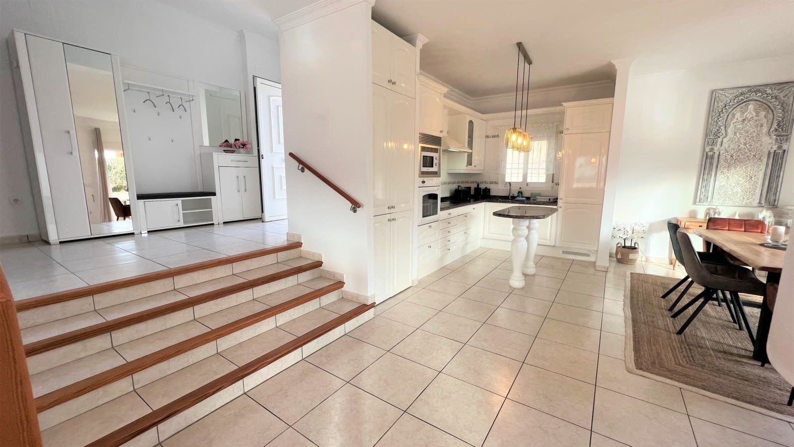 A modern kitchen with white cabinetry, a dining area with a table and chairs, and a staircase leading to another level.