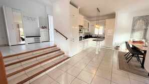 Una cocina moderna con armarios blancos, un comedor con mesa y sillas, y una escalera que lleva a otro nivel.