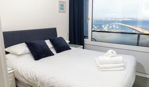 Un lit bien fait, avec du linge de lit blanc et deux oreillers foncés, une serviette blanche posée sur le lit, et une fenêtre donnant sur un port et des bateaux.