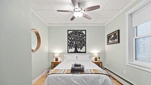 A neatly arranged bedroom with a bed, two bedside tables with lamps, a ceiling fan, a large window, and a wall-mounted artwork. - Beachside Retreat on the Hill (Old Orchard Beach)