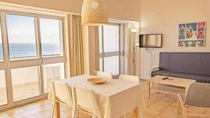 Une pièce comprenant une table à manger, des chaises, un canapé, un téléviseur et de grandes fenêtres donnant sur la mer.