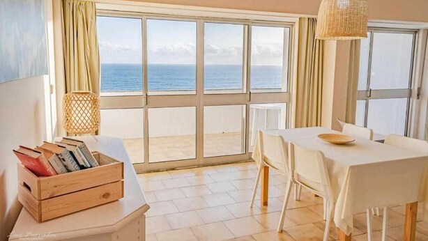 Une pièce dotée d’une grande fenêtre donnant sur la mer, d’une table à manger avec des chaises et d’une lampe en osier.
