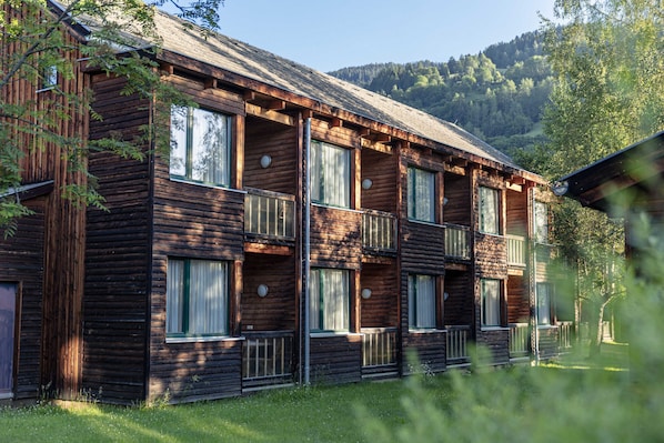 Una cabaña de madera con varios balcones y ventanas amplias, rodeada de follaje y colinas.