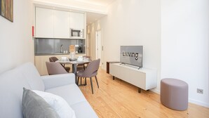 A modern living room with a sofa, a dining table with chairs, a kitchen area with cabinets and appliances, and a television on a stand. - Liiiving-Mouzinho Residence Apartment 1F (Porto)