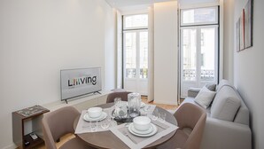 A modern living room with a dining area set for four, featuring a sofa, a TV, and a large window with a view of buildings. - Liiiving-Mouzinho Residence Apartment 1F (Porto)