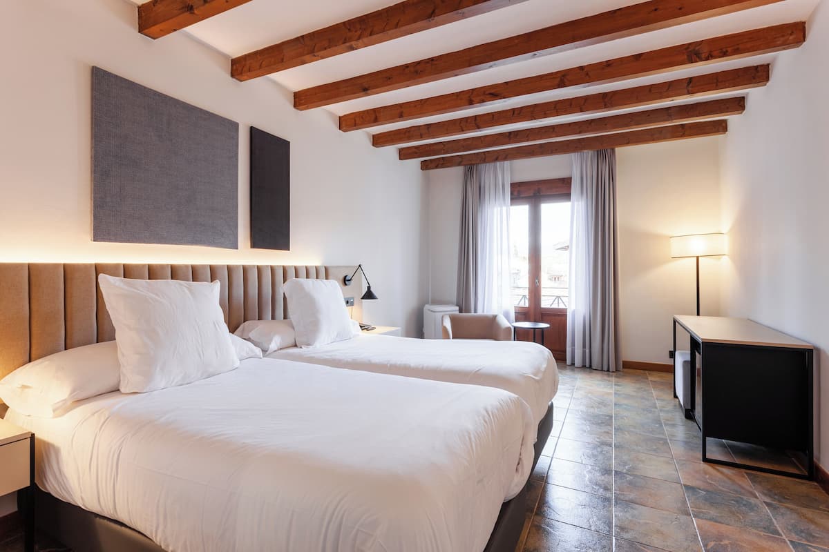 Een hotelkamer met twee bedden, een houten plafond, een bureau, een stoel en een raam met gordijnen.