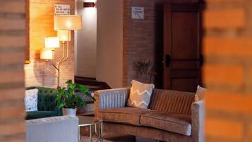 Un hall acogedor con un sofá marrón, un sillón verde y una pequeña mesa con una planta en maceta. Se observan indicaciones hacia el restaurante y la terraza.