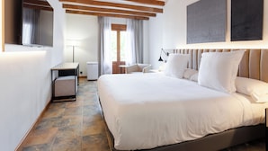 A modern hotel room with a large bed, wooden beams, a flat-screen TV, a desk, and a window with curtains.