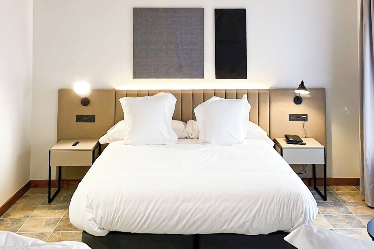 A hotel room with a large bed, two bedside tables, a nightstand, a wall-mounted lamp, and a window with curtains.