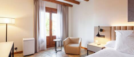 Una habitación de hotel con cama, una silla, escritorio, lámpara, mesita de noche y ventana con cortinas.