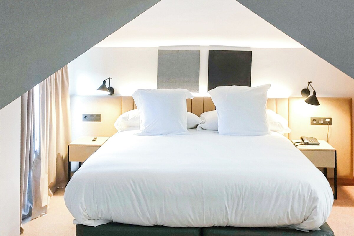 Une chambre d’hôtel avec un grand lit, deux tables de chevet, une petite table, une lampe et une vue sur le ciel.
