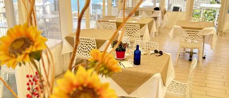 A tented outdoor dining area with tables and chairs, large windows, and a view of the sea.