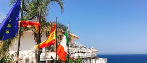 Un edificio costero con varias banderas, entre ellas la bandera de la Unión Europea, y vista al mar.