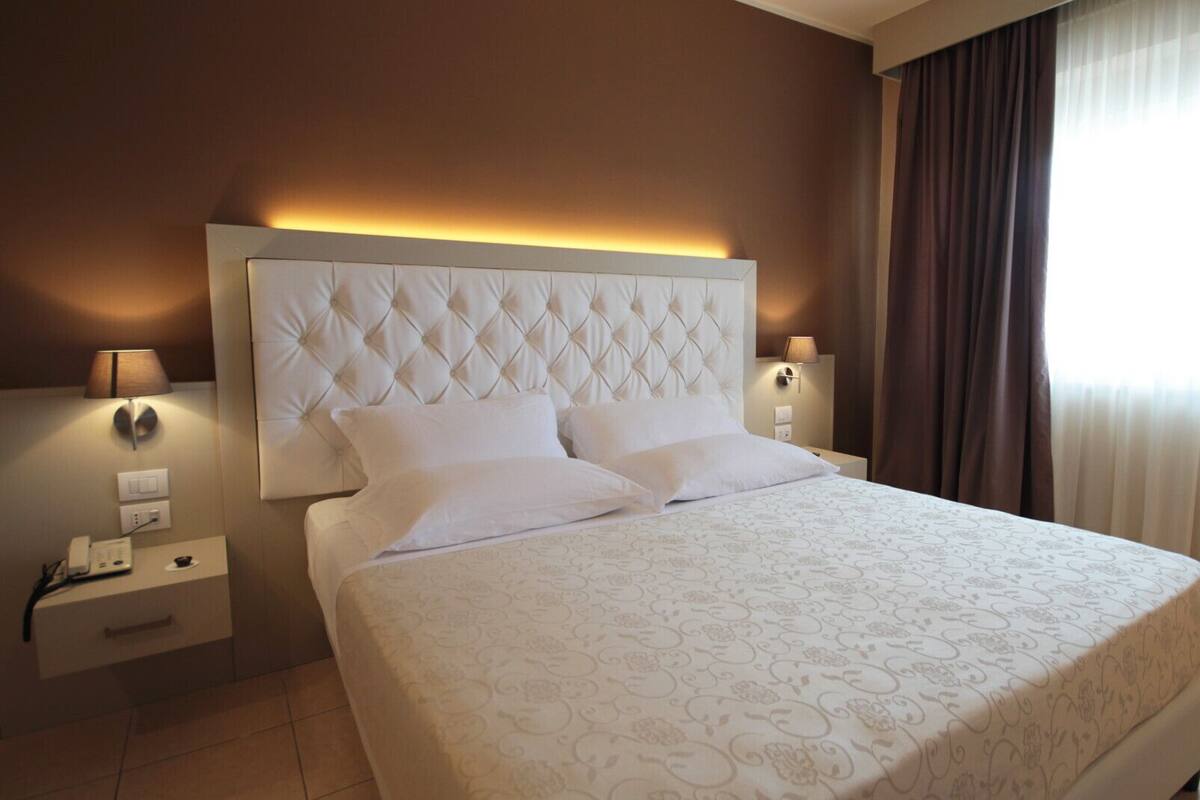 A hotel room with a large bed, two bedside tables, a nightstand, a wall-mounted lamp, and a window with curtains.