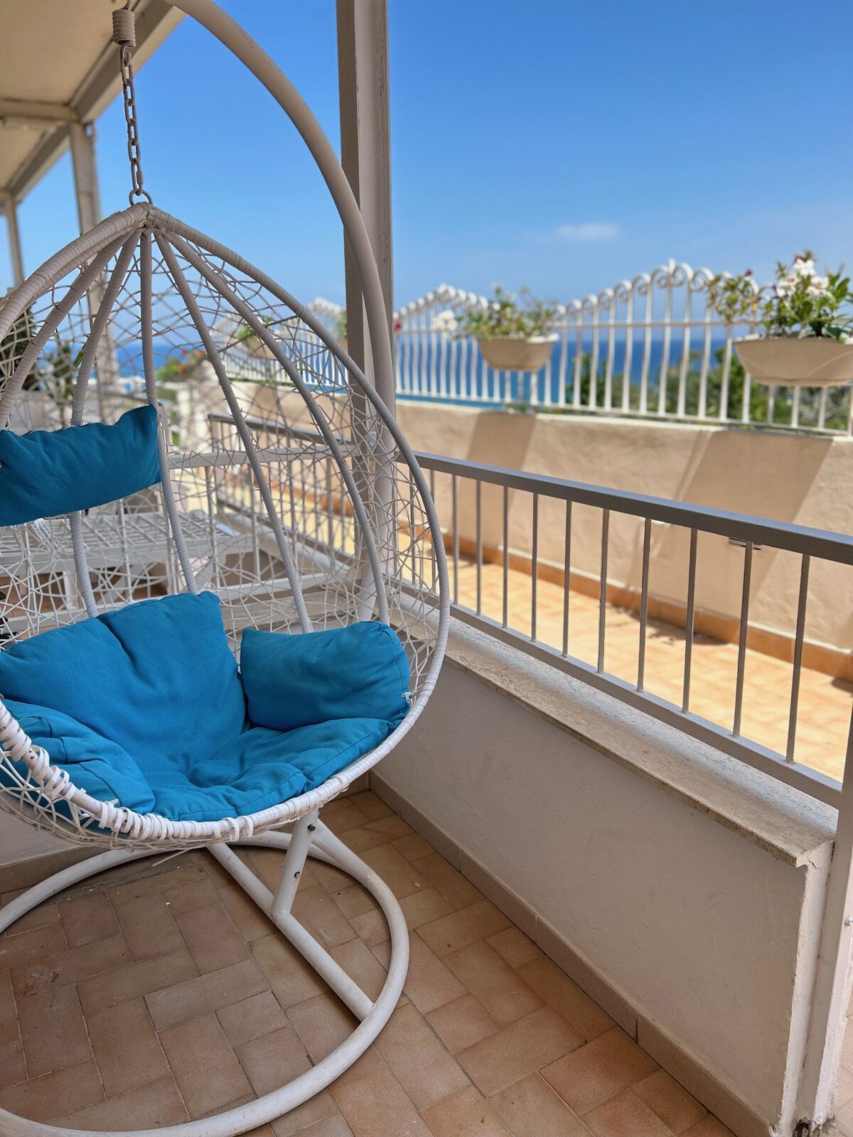 Una silla colgante de mimbre blanco con cojines azules en un balcón con vistas a una valla blanca y vegetación.