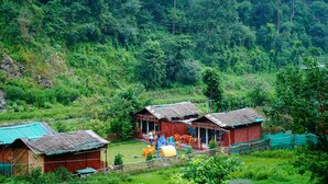 A rural setting with traditional wooden houses, a small outdoor seating area, and a lush green forest in the background. - Kainaat A Riverside Homestay  (Kandaghat)