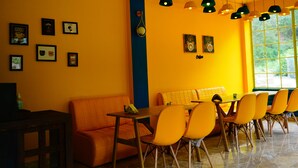 A brightly lit dining area with orange chairs and a table, a blue accent wall, and multiple hanging pendant lights. - Kainaat A Riverside Homestay  (Kandaghat)