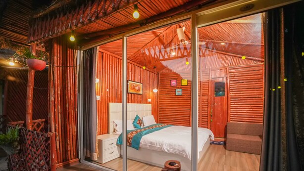 A room with a large bed, a nightstand, a chair, and a sliding glass door leading to an outdoor area with bamboo structures. - Kainaat A Riverside Homestay  (Kandaghat)