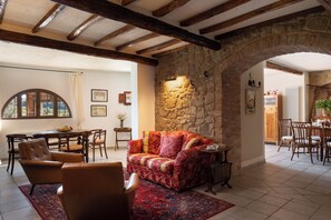 Un salon chaleureux avec un mur en pierre, des poutres apparentes, un canapé rouge à motifs et un coin repas doté de meubles en bois.