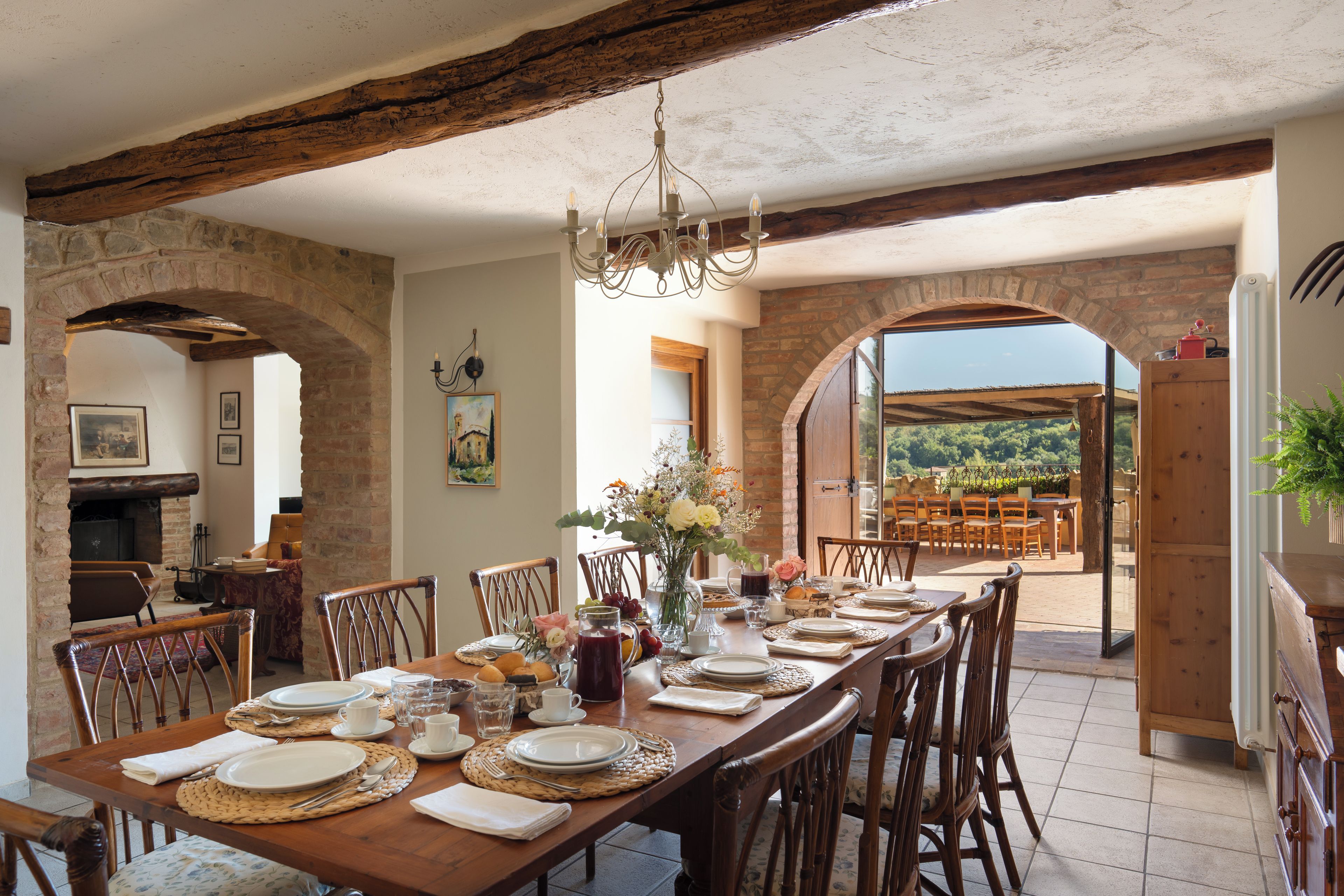 Un comedor con una larga mesa de madera dispuesta para una comida, rodeado de arcos de piedra y vigas de madera.