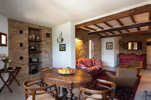 A traditional room with exposed brick walls, a wooden ceiling, a round wooden table with chairs, a red patterned sofa, and a stone fireplace.