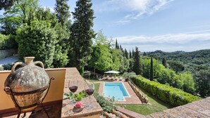 A patio with a large ceramic pot and wine glasses, overlooking a swimming pool and lush greenery.