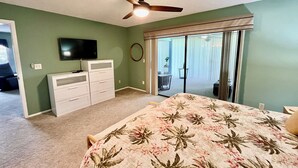 Un dormitorio con cama, ventilador de techo, televisor, cómoda y una puerta corrediza de vidrio que da a un balcón.