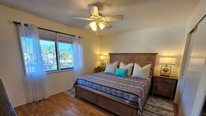 Un dormitorio con cama de madera, ventilador de techo, mesita de noche con lámpara y vistas a palmeras a través de la ventana.