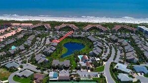 Vista aérea de un complejo turístico con una fuente circular central, rodeada de edificios de estilo residencial y una playa al fondo.