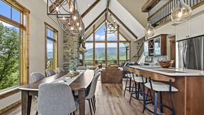 Ein geräumiger Essbereich mit einem langen Holztisch und Stühlen, einem Kamin aus Stein und großen Fenstern mit Blick auf die Berge.