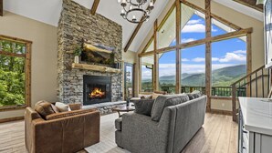 Ein großzügiges Wohnzimmer mit Kamin aus Stein, großen Fenstern und Blick auf die Berge.