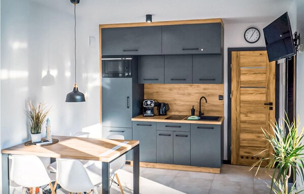 A modern kitchen with grey cabinets, a wooden countertop, and a coffee machine. There is a dining table with chairs and a hanging pendant light.