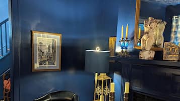 Una habitación con pared azul oscuro, un cuadro enmarcado, una lámpara, un candelabro, una silla, una alfombra y una chimenea.